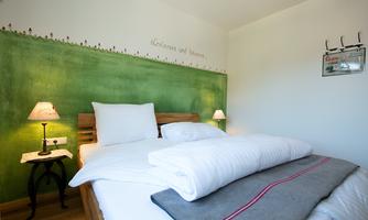 A cozy bedroom with a green accent wall. The bed is neatly decorated with white and gray bedspreads and pillows.