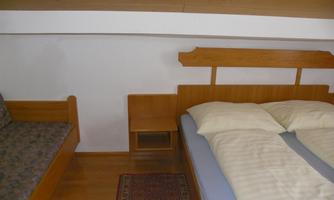 A simple bedroom with a double bed and a small couch. The walls are light, and the floor is made of wood.