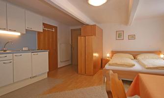 A cozy room with a double bed and a small kitchenette. The floor is made of wood and there is a dining table with chairs.