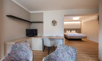 A modern hotel room view with a desk and two cozy armchairs. In the background, a bedroom with a large bed is visible.