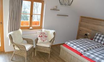A cozy bedroom with a double bed and two wicker chairs. Large windows let in plenty of light and offer a view outside.