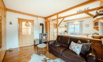 A cozy living room with a brown sofa and a wood stove. The inviting atmosphere is complemented by wooden walls and an open kitchen.
