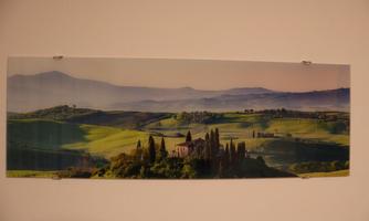A picturesque landscape with gentle hills and wide green fields. In the foreground stands a historic building surrounded by cypress trees.
