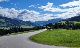 A winding road leads through a green landscape with mountains in the background. The sky is clear and blue with a few clouds.
