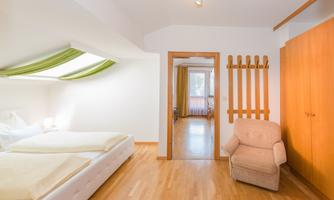 A bright bedroom with a double bed and a comfortable armchair. In the background, an entrance area is visible.