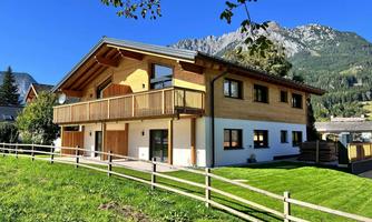 A modern wooden house with a balcony and large windows. It is situated in a green environment with mountains in the background.