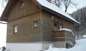 A cozy wooden house in the snow, surrounded by a wintry landscape. The snow-covered facade and the rustic style create an inviting atmosphere.