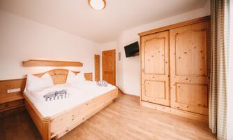A cozy room with a double bed and wooden furniture.  
A television is hanging on the wall, and the light is bright and inviting.