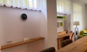 A bright room with a wooden log table and large windows. On the wall, there is a clock and some shelves with green accents.