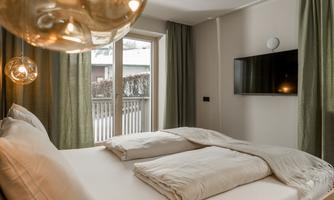 A modern bedroom with a large bed and white bedding. Large windows with green curtains provide plenty of daylight.