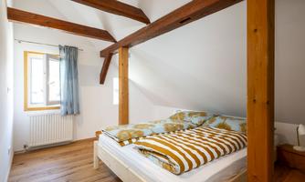 A cozy bedroom with wooden flooring and a sloped ceiling. The bed is decorated with colorful blankets, and a window lets in plenty of daylight.