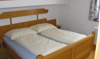 A simple bedroom with a wooden double bed and a small rug. The room has a sloping ceiling and a window with curtains.