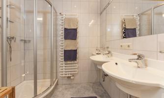 A modern bathroom with a shower cabin and two sinks. The walls are tiled and towels are neatly hung up.