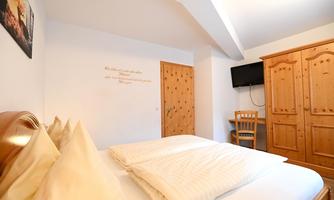 A cozy bedroom with a double bed and bright bedding. In the background, there is a wooden furniture piece and a television on the wall.
