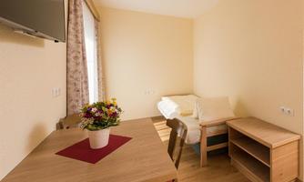 A simple room with a table, a chair, and a television on the wall. A bed and a small flower vase give the room a cozy atmosphere.