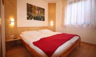 A cozy bedroom with a large bed and a red blanket. On the wall hangs a picture of a forest landscape, and the room has large windows with curtains.