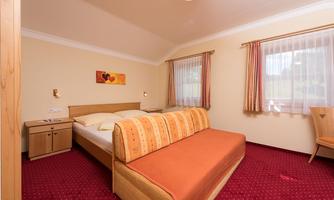 A cozy bedroom with a double bed and a seating area. The walls are painted light, and there are large windows with curtains.