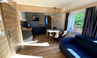 A cozy living space with a blue sofa and a dining table. The surroundings are bright and modern, featuring wood decor and large windows.