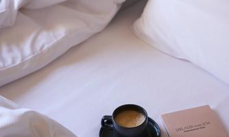 A cozy bed with white pillows and a blanket. A cup of coffee and a book are lying on it.