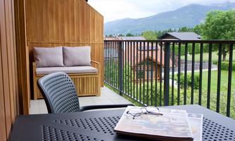 A balcony with a table and a chair. In the background, green meadows and mountains can be seen.