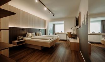 A modern bedroom with a double bed, wooden flooring, and clean lines. There are windows facing outside and a cozy corner with a desk.