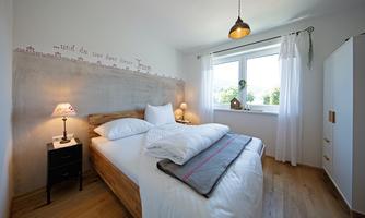 A cozy bedroom with a large bed and a window that lets in plenty of light. The walls are bright, and there is a decorative lamp as well as a small bedside table.
