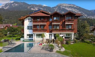 A charming multi-story house with a balcony, surrounded by mountains. In the foreground, there is a pool and a green lawn area.