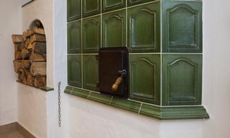 A traditional tiled stove with green tiles and a wood storage area. Next to it is a black oven door with a wooden handle.