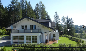 A modern house surrounded by green meadows and tall trees. The clear sky and quiet landscape create an idyllic atmosphere.