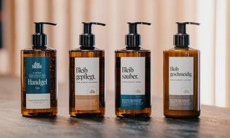 Four bottles of care products are placed on a wooden table. The bottles have attractive labels with various texts, including "Hand gel," "Stay cared for," "Stay clean," and "Stay smooth."