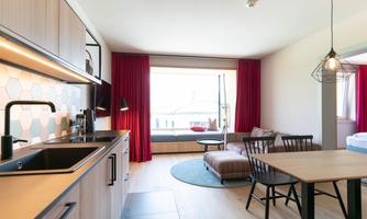 A modern kitchen and living area with bright furniture and large windows. Red curtains and a round rug give the room a cozy atmosphere.
