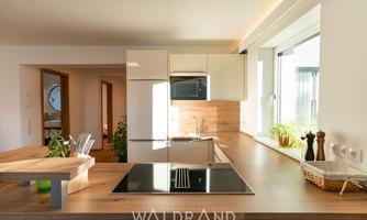 A modern kitchen with wooden surfaces and bright walls. The large windows provide plenty of daylight and a welcoming atmosphere.