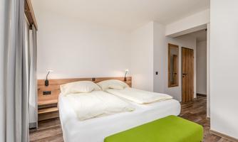 A modern bedroom with two single beds and a green stool. The walls are bright, and the room feels spacious and inviting.