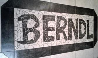 A creative mosaic design named "BERNDL" on a floor. The pattern consists of various stone fragments and has a modern, stylish frame.