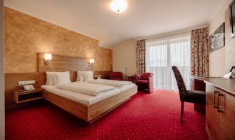 A cozy hotel room with two beds, a desk, and a comfortable armchair. Large windows let in plenty of light and offer a view of the outdoor area.