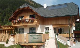 A charming wooden house with flower balconies and a friendly entrance. The surroundings are enveloped by green mountains.