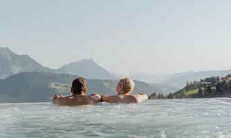 A couple relaxes in an outdoor hot tub with a view of the mountains. The scenery displays a breathtaking landscape and a clear sky.