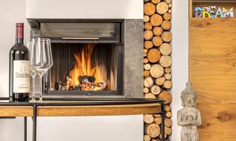A cozy fireplace with a crackling fire and a table on which a bottle of wine and two glasses are placed. Next to the fireplace are stacked logs and a decorative statue.