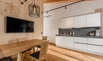 A modern kitchen with a wooden table and chairs. A television is hanging on the wall, and there is an elegant pendant light.