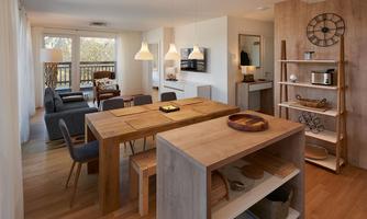 A modern living room with a large wooden dining table. Light streams through large windows, creating an inviting atmosphere.