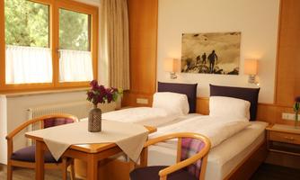 A cozy room with two beds and a small table. The windows let in a lot of light and the walls are decorated with nature-themed pictures.