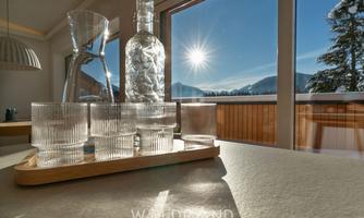 A stylish presentation of glasses and a carafe set on a table. In the background, the sun shines over the mountains and through large windows.