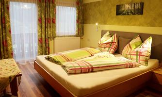 A cozy bedroom with a large bed and colorful pillows. The windows are decorated with blooming curtains, and there is a warm wooden floor.