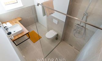 A modern bathroom with a glass shower and a minimalist decor. A wooden shelf with towels and toiletries complements the simple design.