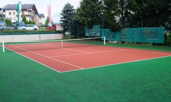 A tennis court with a red surface and green surroundings. In the background, there are trees and some buildings visible.