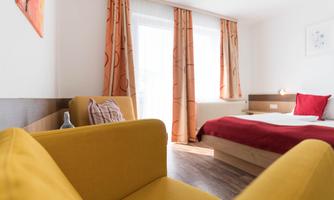 A bright room with a red bed and a yellow armchair. Large windows let in plenty of daylight.