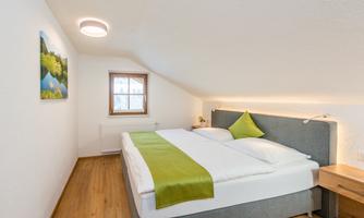A cozy bedroom with a double bed and a green blanket. The room has a sloping ceiling and a window with a view of nature.