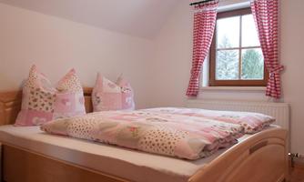 A cozy bedroom with a wooden bed and colorful pillows. The windows are decorated with checked curtains and offer a view of the outdoors.