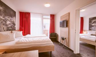 A cozy hotel room with two beds and red curtains. The room is bright and modernly furnished, with a television and a window to the terrace.