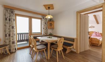 A cozy dining area with wooden chairs and a table in a bright apartment. The windows offer a view of the balcony and allow plenty of daylight.